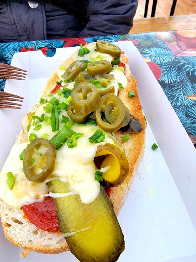 An open faced sandwich on toasted bread with tomato, melted raclette cheese, a huge slab of pickle, jalapeños, green onions and tomatoes