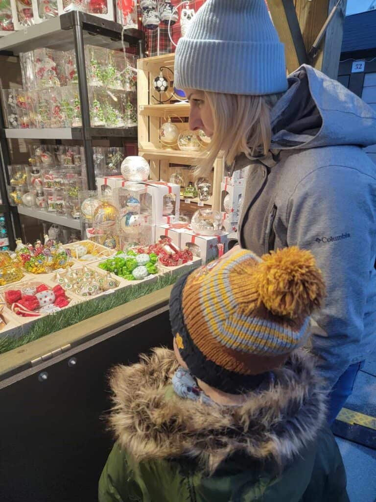 A blonde woman in a blue hat looks at Christmas treats in a booth with her young son who wears a winter coat and a  brown hat with a pom pom