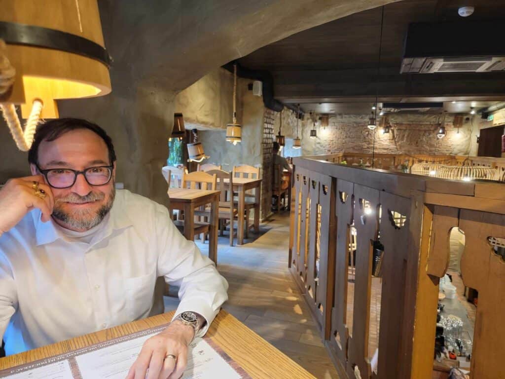 A man smiling at the camera sits at a wooden table on the upper level of a restaurant with stone and wood interior