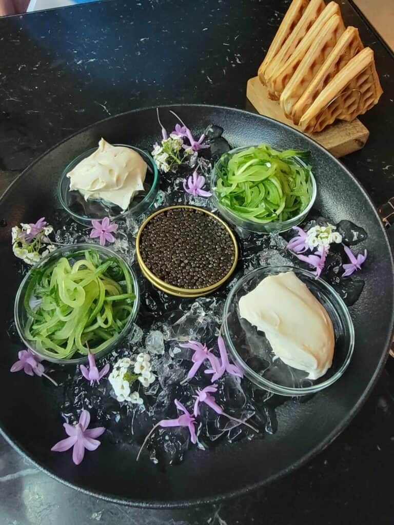 A plate decorated with edible flowers, julienne cucumbers, creamed cheese, and a tin of caviar in the center