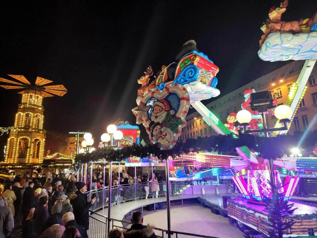 A Christmas-themed carnival ride, crowds of people, and a lit Christmas pyramid in the background