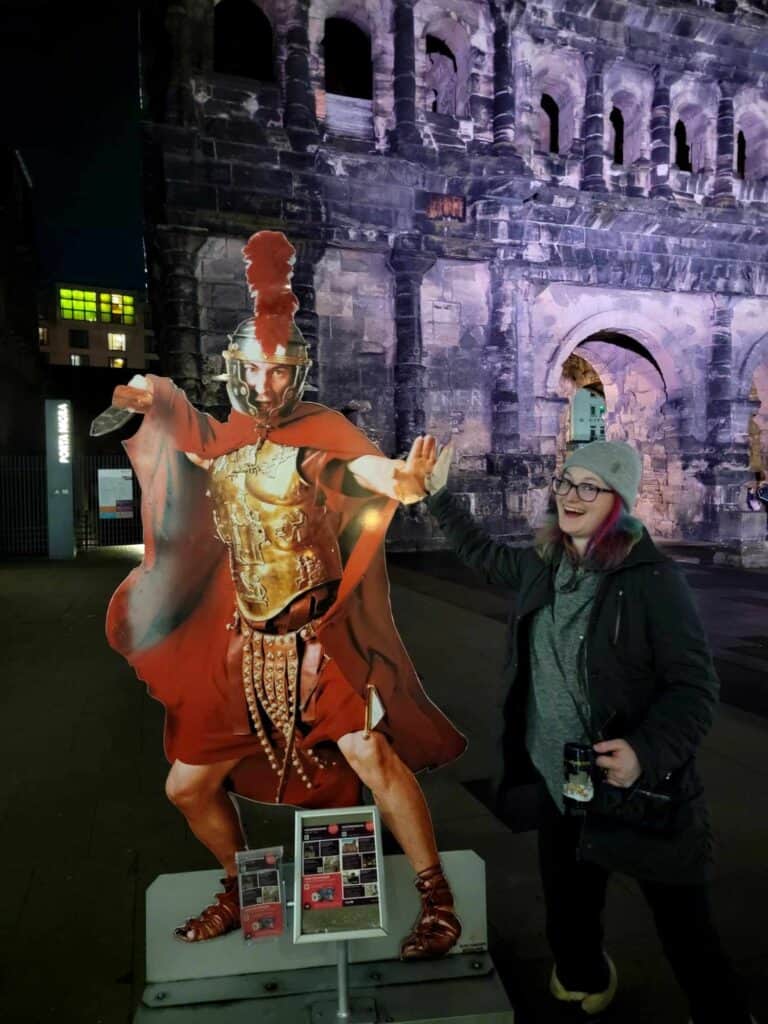 A woman in a knit hat holding a Christmas mug high fives a life-sized cutout of a Roman legionnaire with the Porta Nigra in the background