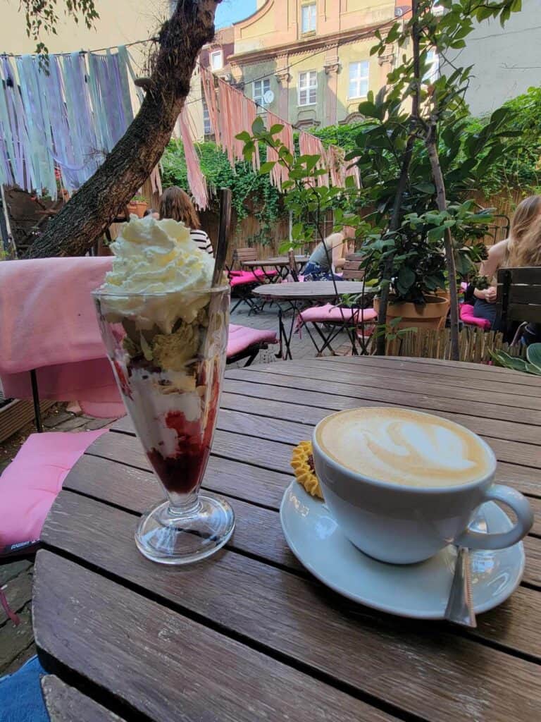 A cappuccino and a tall ice cream sunday on a wooden table in a garden with tables and chairs with pink cushions