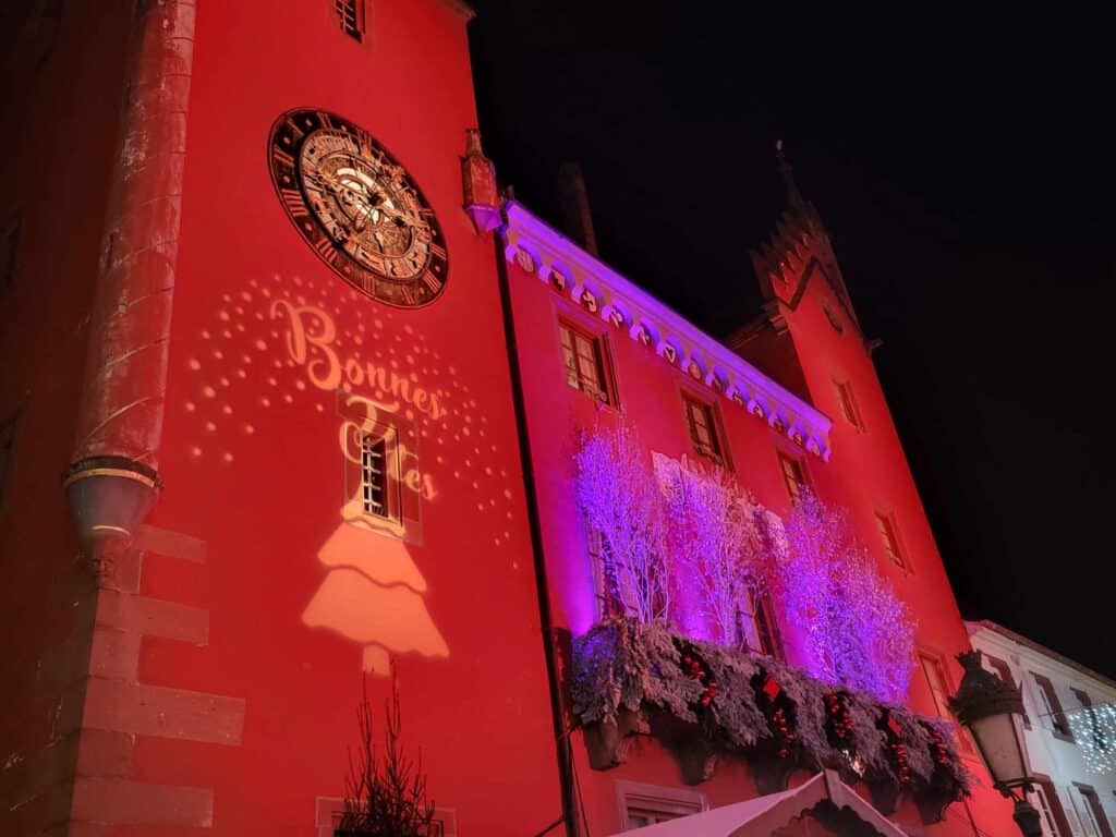 A building illuminated in red and purple with an old clock and the words Bonnes Fetes and a christmas tree