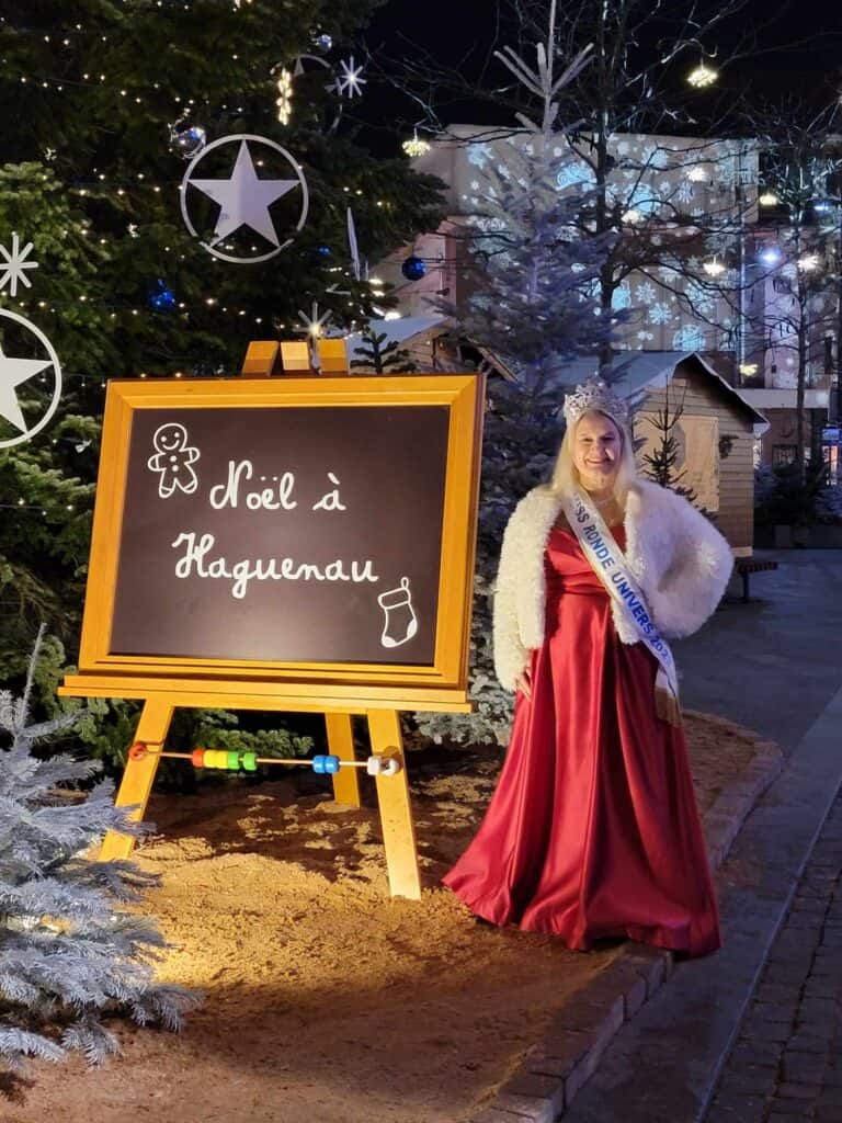 A blonde woman in a red dress and fuzzy white coat wears a pageant sash and stands by a sign that says Noël à Haguenau