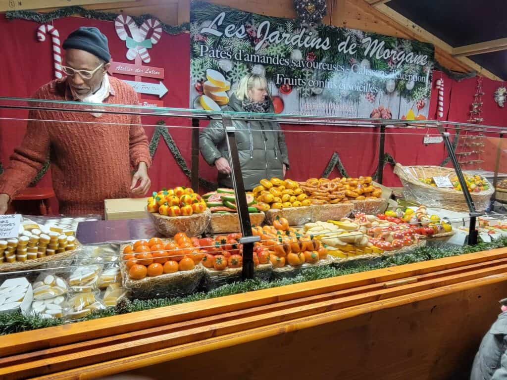 A woman stands behind the counter of a booth selling marzipan shaped like fruits and pretzels