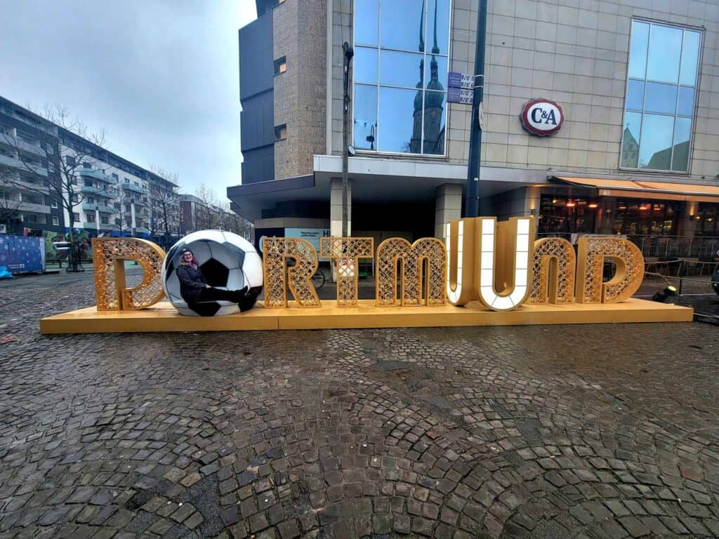 A lit sign on the street that says DORTMUND in large illuminated letter. The O is a soccer ball and a woman sits inside it smiling