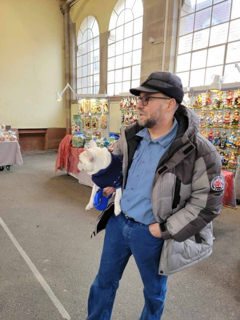 A man holds a white frenchie like a football in front of indoor market stalls