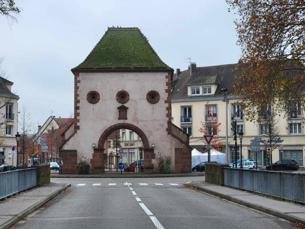 A white medieval tower with a flat topped green roof, three small round windows, over an arch framed in red sandstone in the center of a traffic island