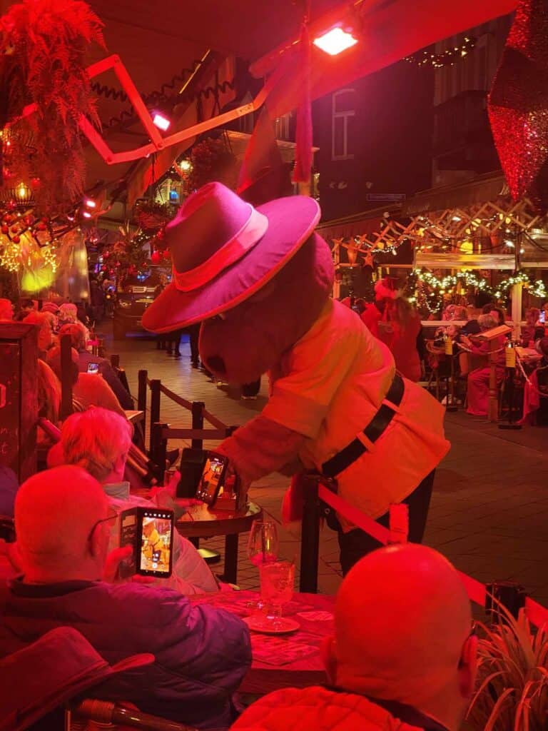 A large bear in a fedora leaning over a rail, greeing a woman sitting at a cafe table bathed in red light