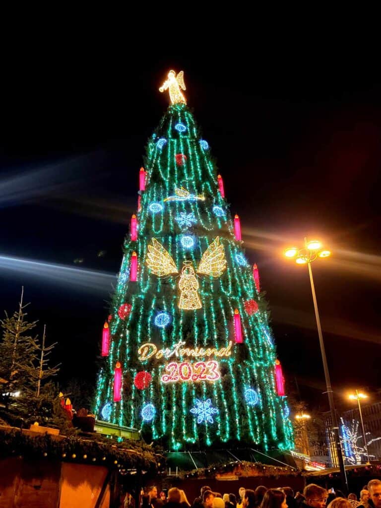 A huge Christmas tree with a golden angel on top and market stalls below. It says Dortmund 2023