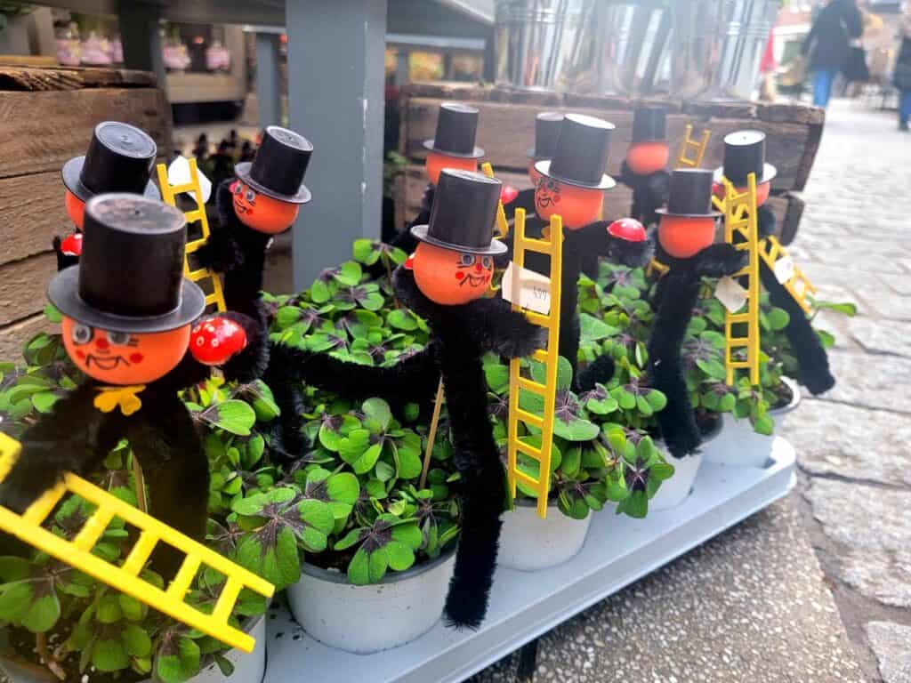 Chimney sweeps made of pipecleaners with round heads in tophats holding small yellow paper ladders, red mushrooms, and next to a small bit of potted clover