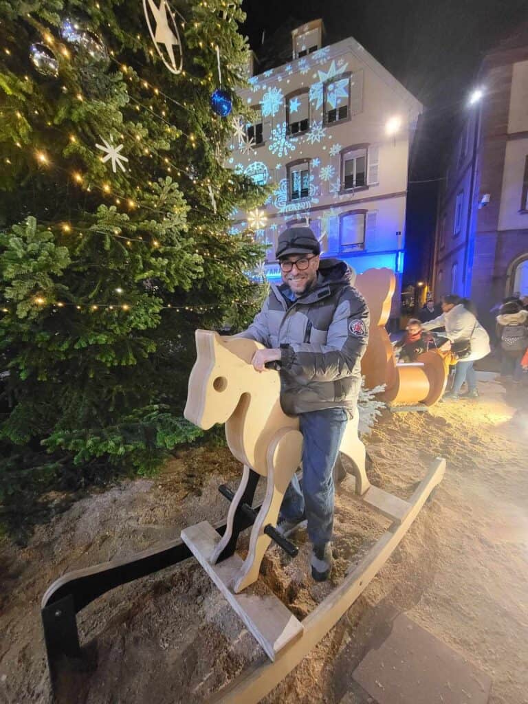 A man in a winter jacket sits astride a large wooden rocking horse next to a Christmas tree