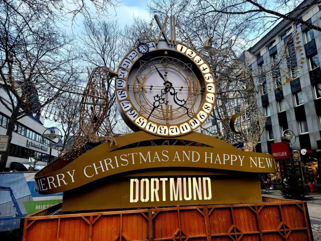 An old fashioned clock with numbers around it from 1 to 24. They are lit up to and including 15. The sign below says Merry Christmas and Happy New Year Dortmund