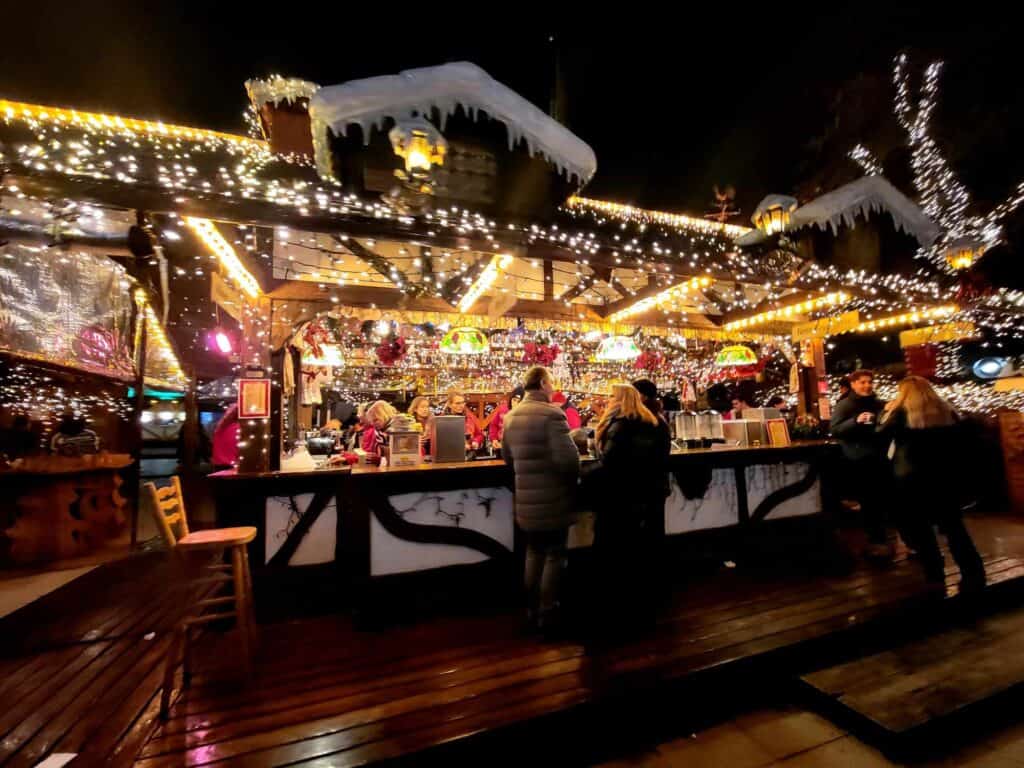 A wooden market stall decorated in icicles and little white lights. People stand in front with mugs talking