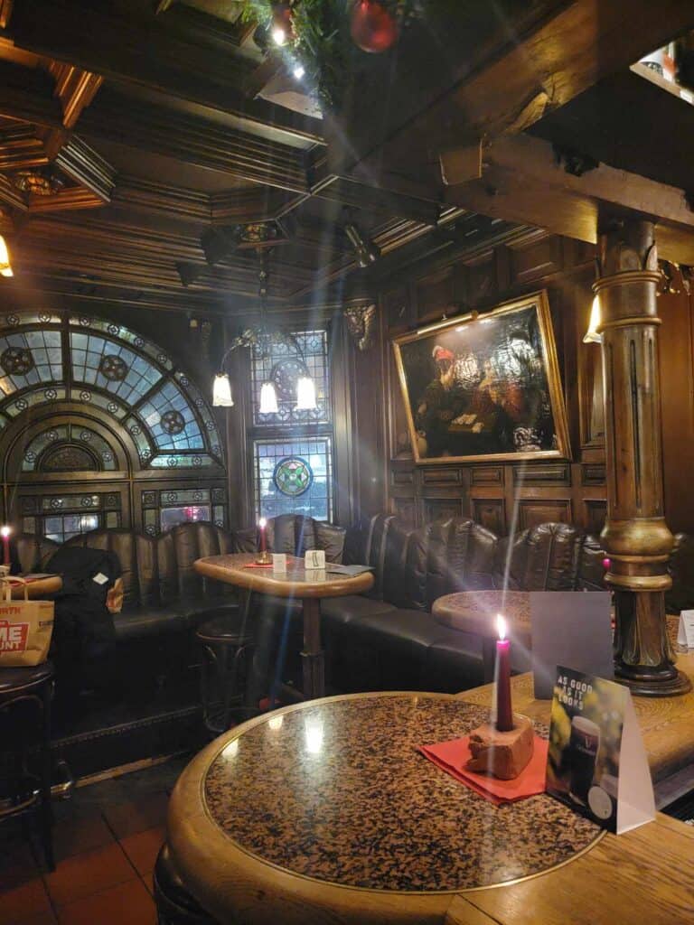 The inside of a pub. Everything is wooden and dark. There are leaded windows, old paintings, and marble table tops with candles