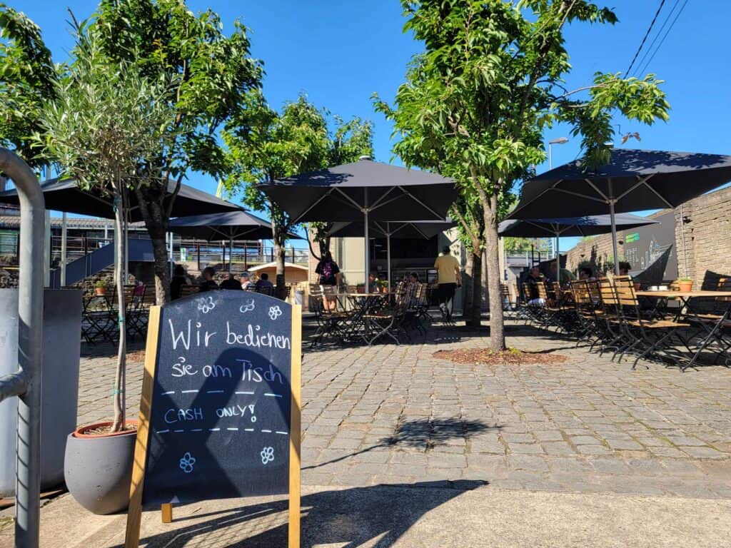 A biergarten with trees and a chalkboard sign advertising prices. Tables and umbrellas behind