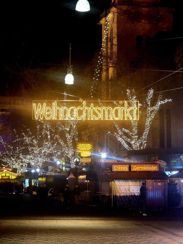 A large illuminated sign over a street saying Weihnachtsmarkt and a line of trees covered in little white lights, and a booth that says Crêpes, Germknodel.