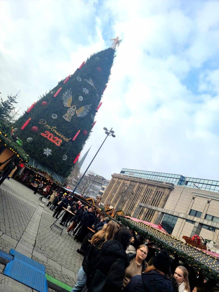 A huge Christmas tree with market stalls in front and blue sky with clouds. A sign on the tree says Dortmund 2023