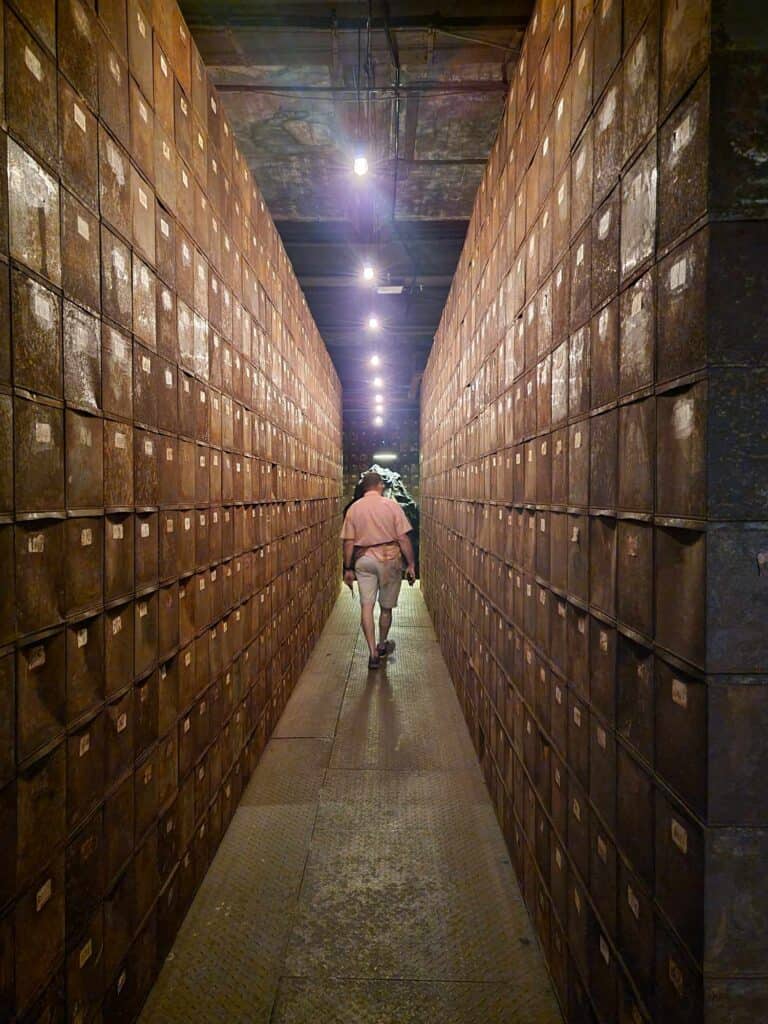 A man in tan shorts and a pink shirt walks down a narrow alley made of tall walls of brown boxes with numbers