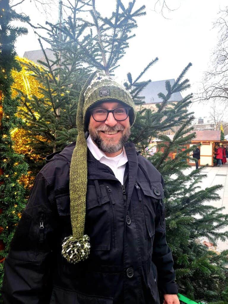 A man in glasses smiling wearing a long green knit hat with a pom pom