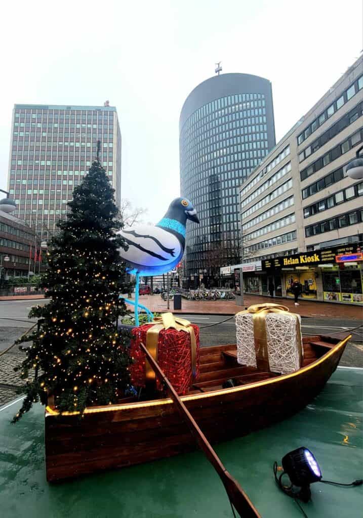 A pigeon with long flamingo legs sitting in a huge rowboat with presents and a Christmas tree