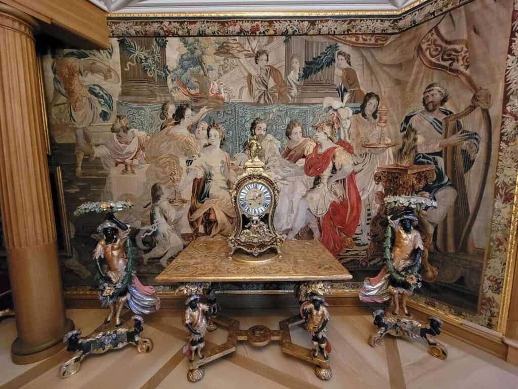 A gilt table with an ornate clock in front of a Belgian tapestry