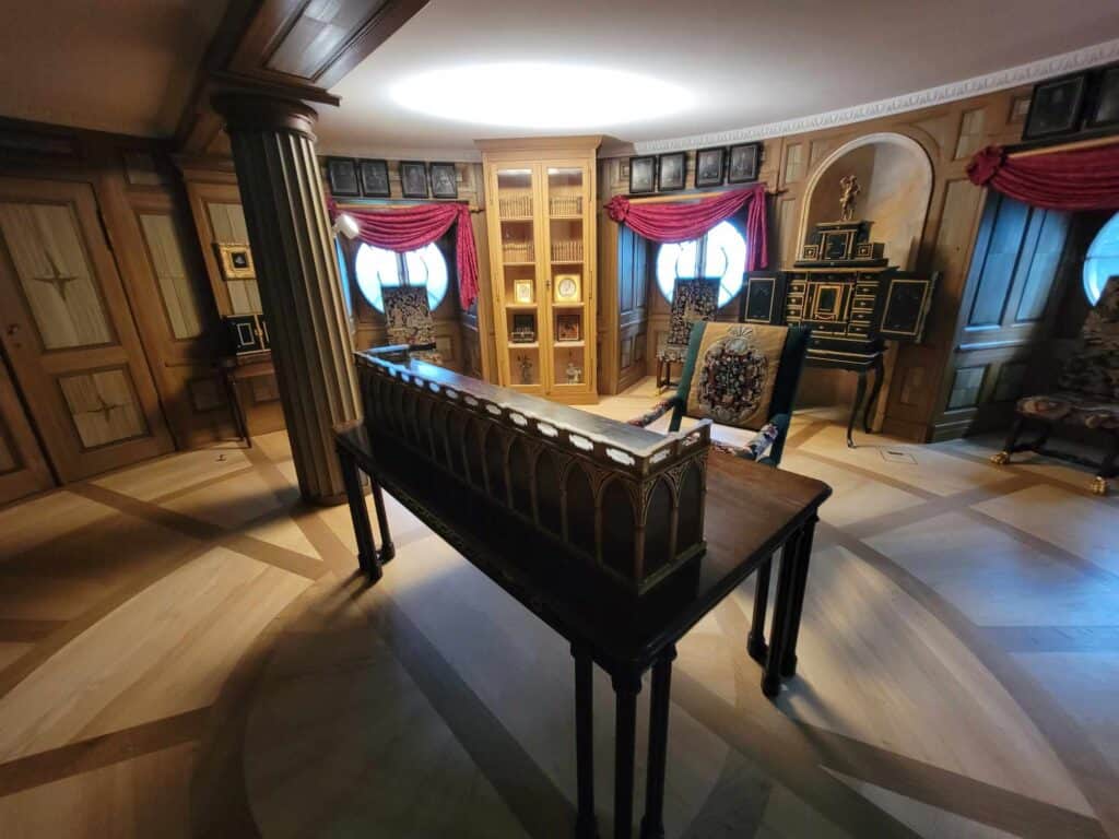 A well-appointed library with wooden shelves, a desk in the centre and red draperies