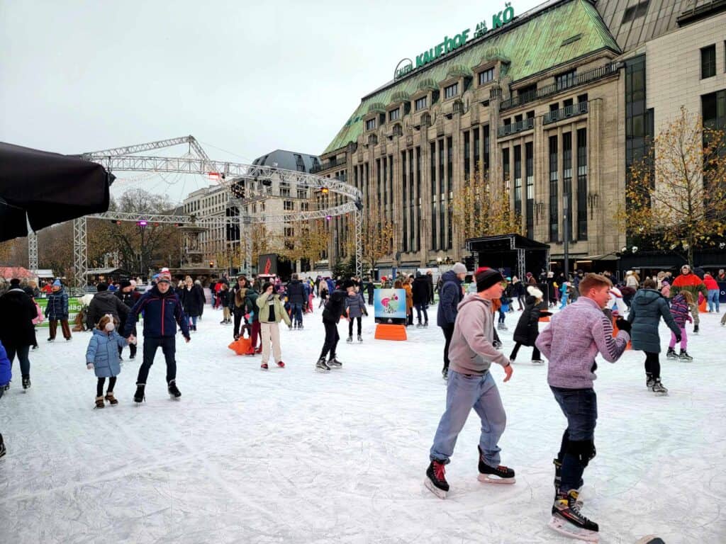 A large ice rink in the city with many people of all ages skating