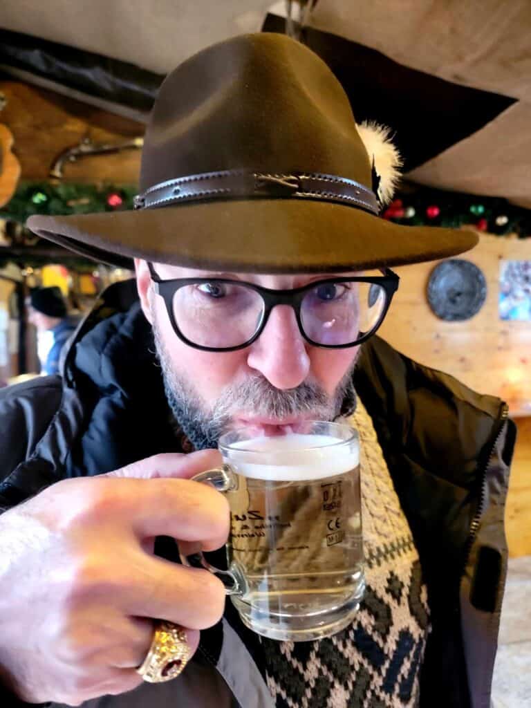 A man in a ahat sipping hot white gluhwein out of a clear glass mug with writing on it