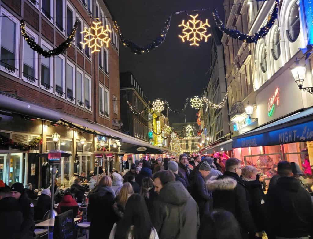 A busy street full of people with shops on either side and illuminated snowflake decorations above