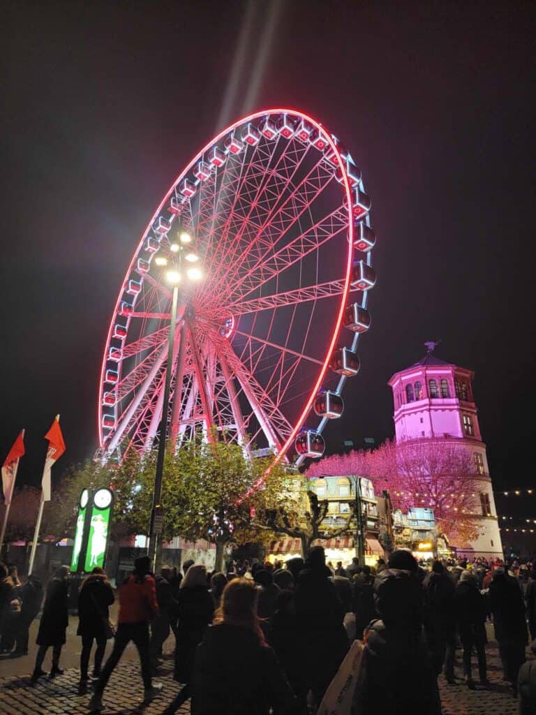 An illuminated ferris wheel in pink and blue next to a medieval tower