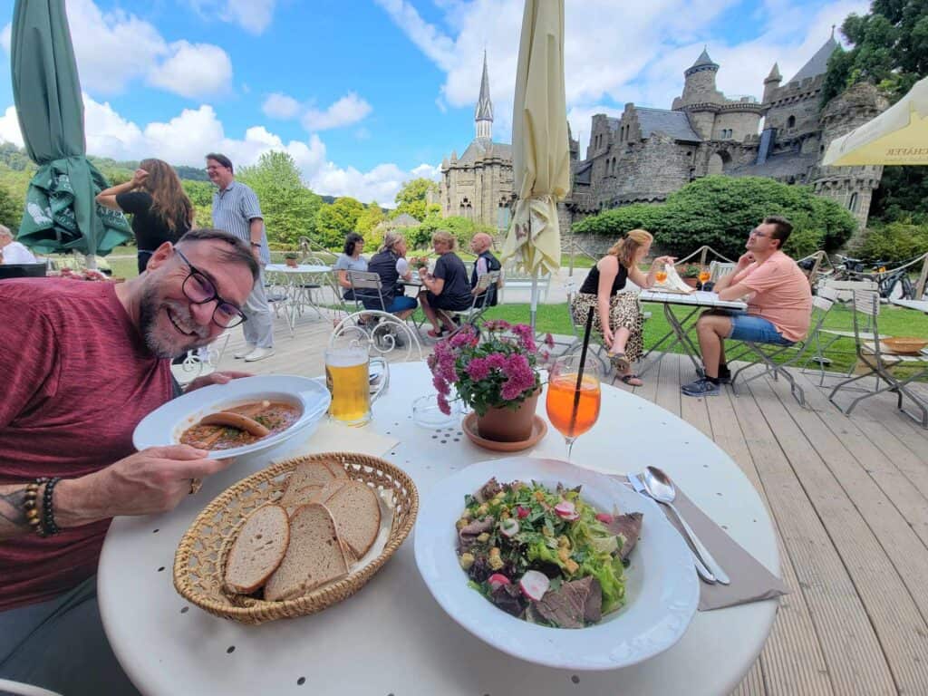 A smiling man holding a plate of lentils and sausages with a gorgeous castle behind