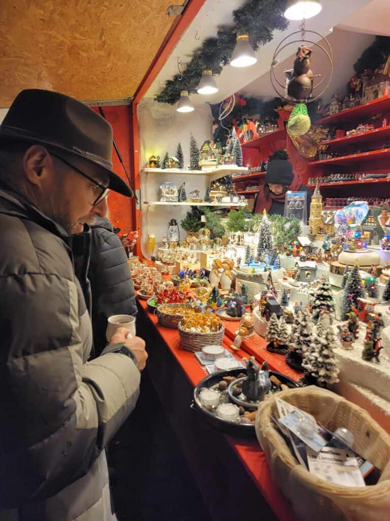 A man in a hat with a mug of gluhwein looking at a booth full of Christmas decorations at the Dusseldorf Christmas market