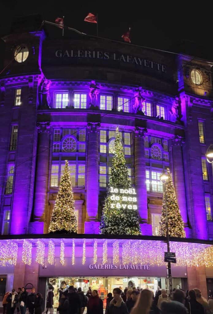 The Galeries Lafayette illuminated in purple with hanging icicle lights and tall Christmas trees with a sign saying "le noel de mes reves"