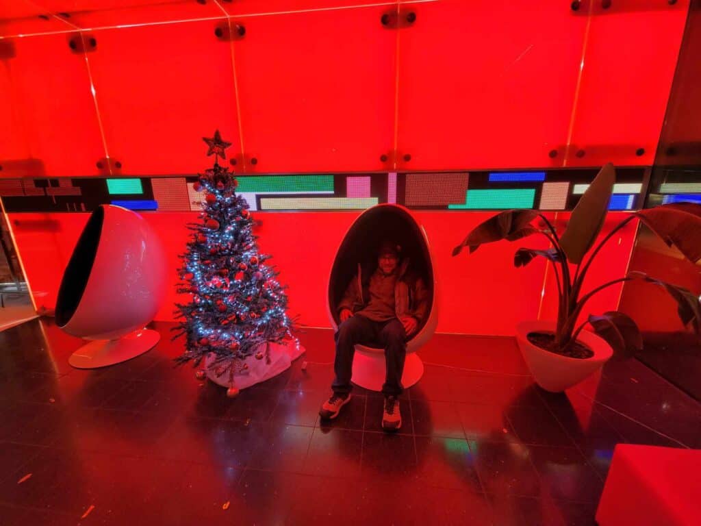 A man in a hotel lobby flooded with red light sitting in a chair shaped like an egg next to a Christmas tree