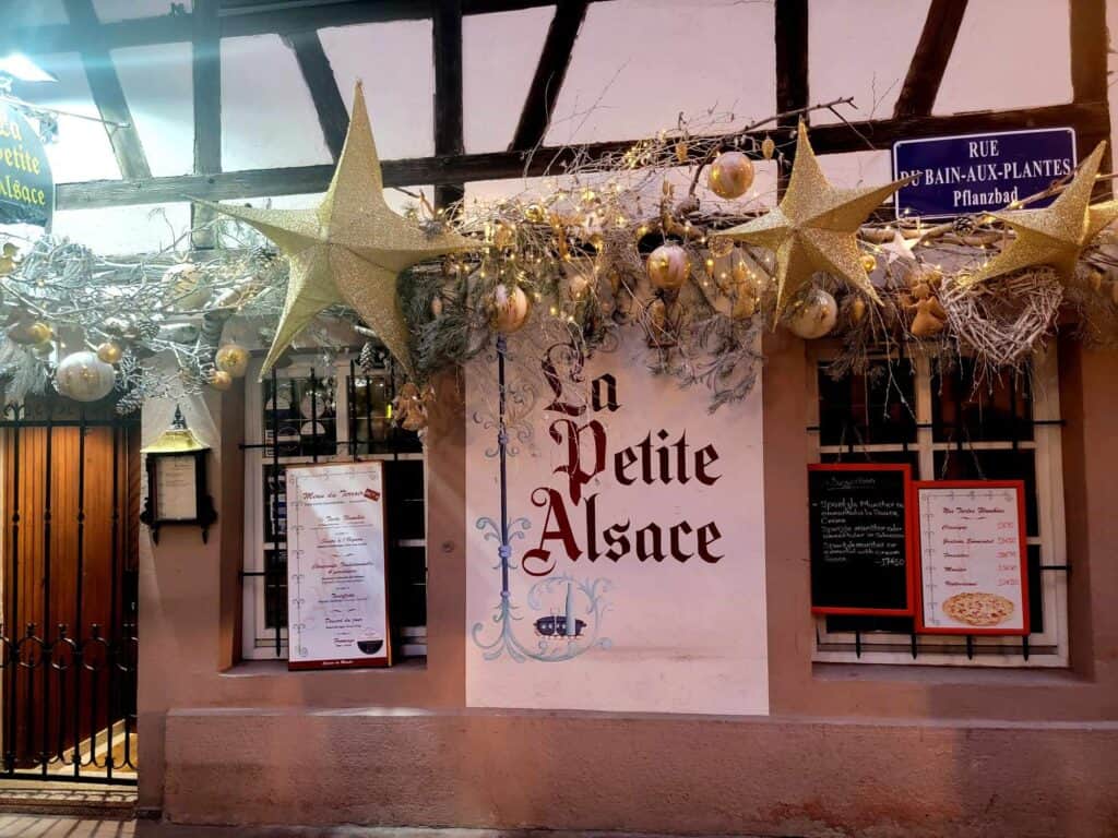 A sign on a half timbered building saying La Petite Alsace with a garland covered with golden ornaments