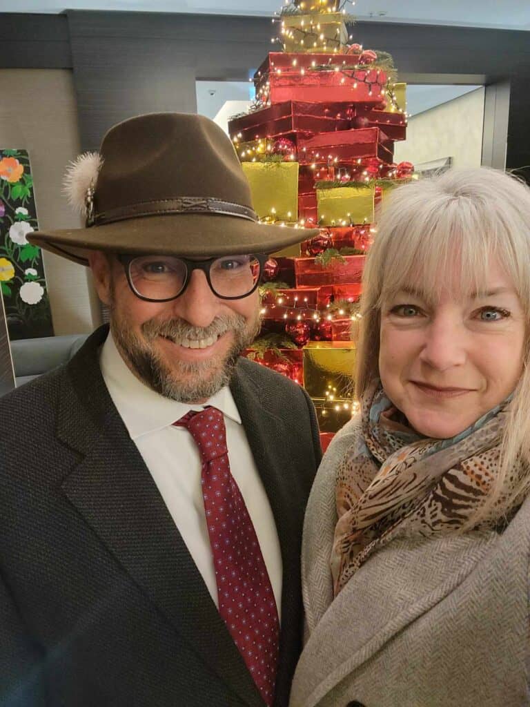 A smiling couple dressed to go out with a large pile of red and gold boxes covered with Christmas lights behind.