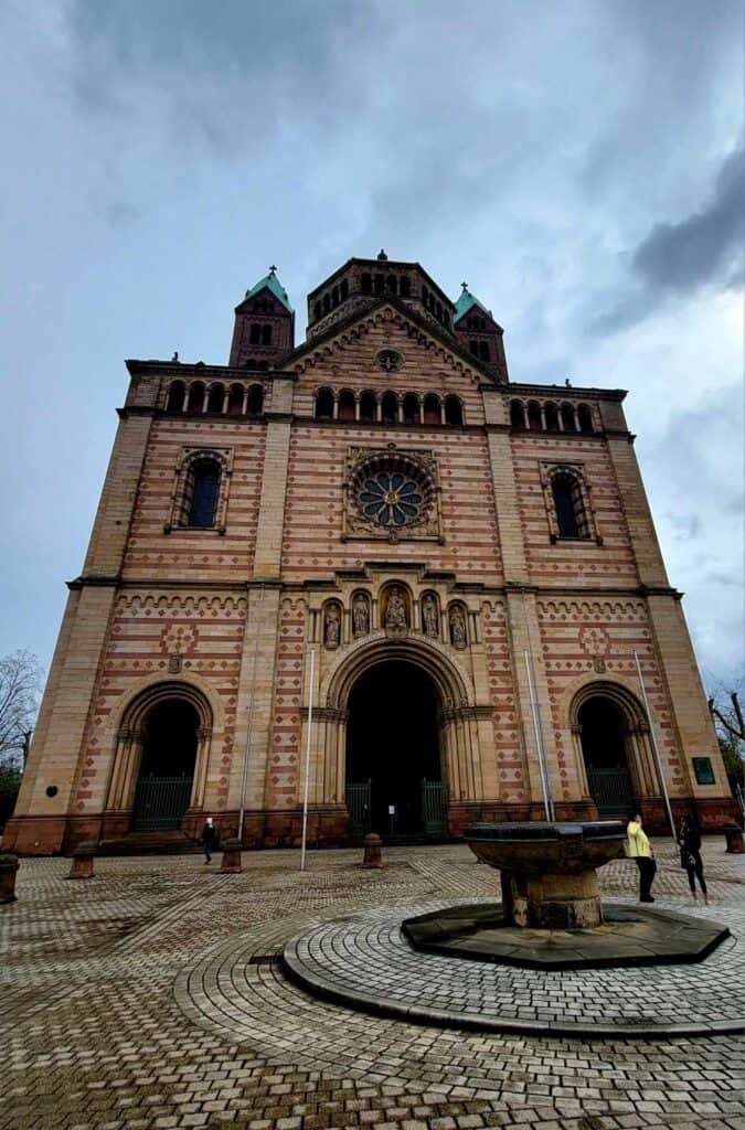 The two-toned front of the Speyer cathedral with an arched main door and two arches on the sides, and two towers in the background