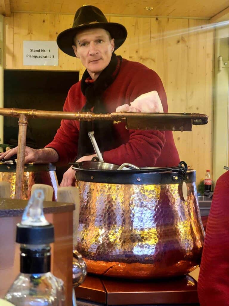 A man in a hat behind a copper kettle full of gluhwein