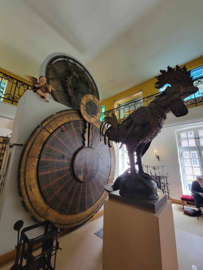 A large old bronze rooster stands in front of an ancient clock dial