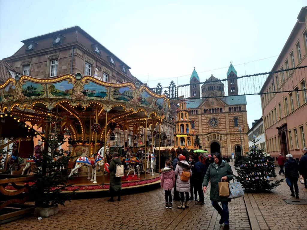 An antique carousel with horses surrounded by Christmas trees and people. Speyer cathedral behind.