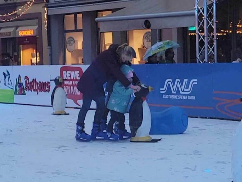 A woman standing behind a child on ice skates holding a penguin on skis to stabilize herself