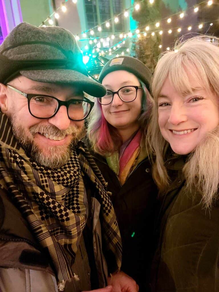 Three smiling people in coats with Christmas lights behind