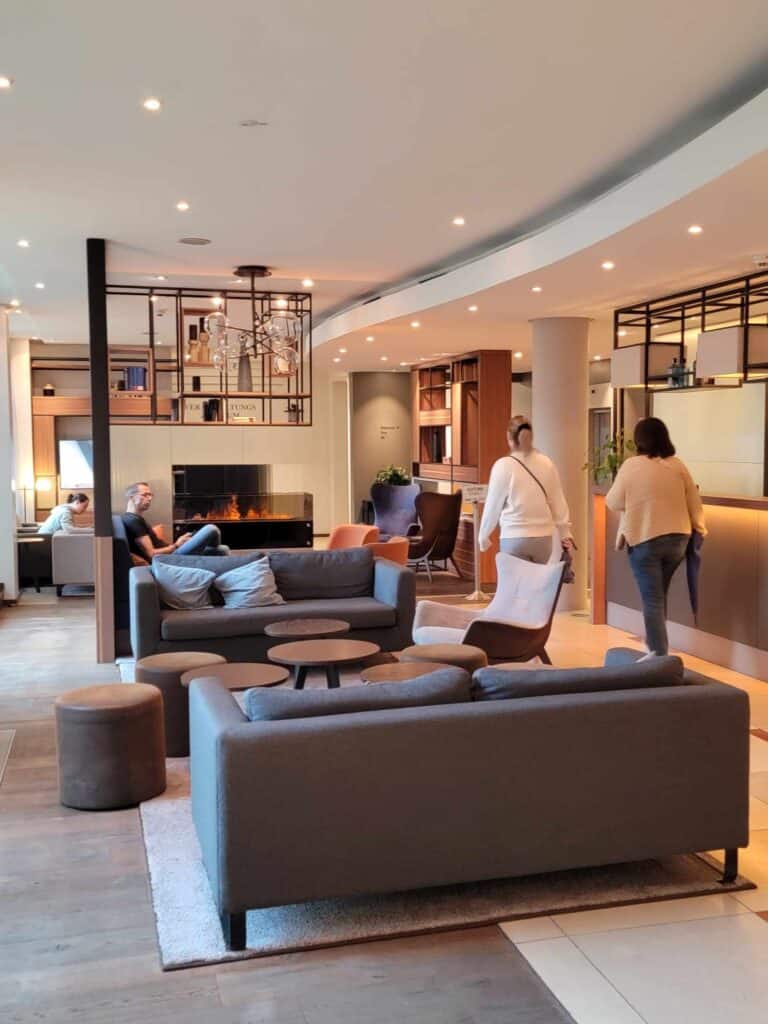 A hotel lobby with grey love seats and upholstered stools, with tables and a small gas fireplace with people sitting and walking