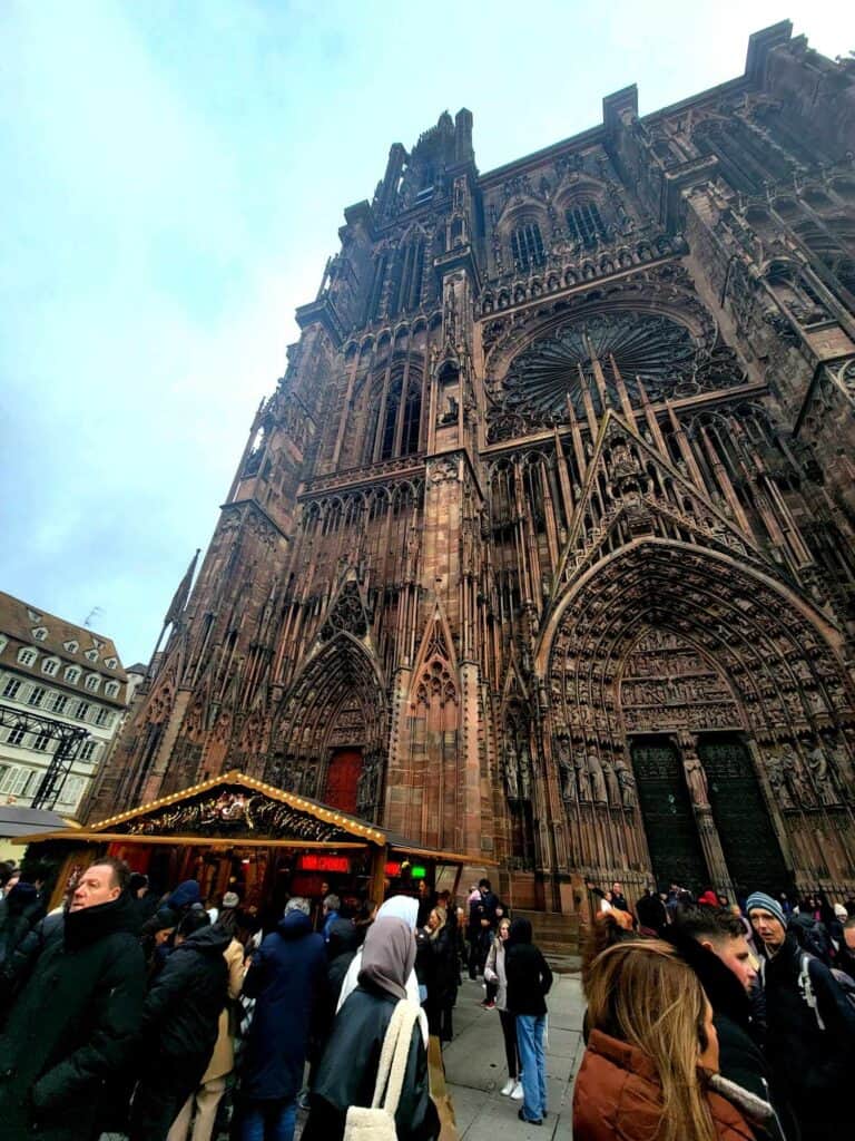 The facade of Strasbourg Cathedral with heavily detailed gothic ornamentation and people shopping at market stalls