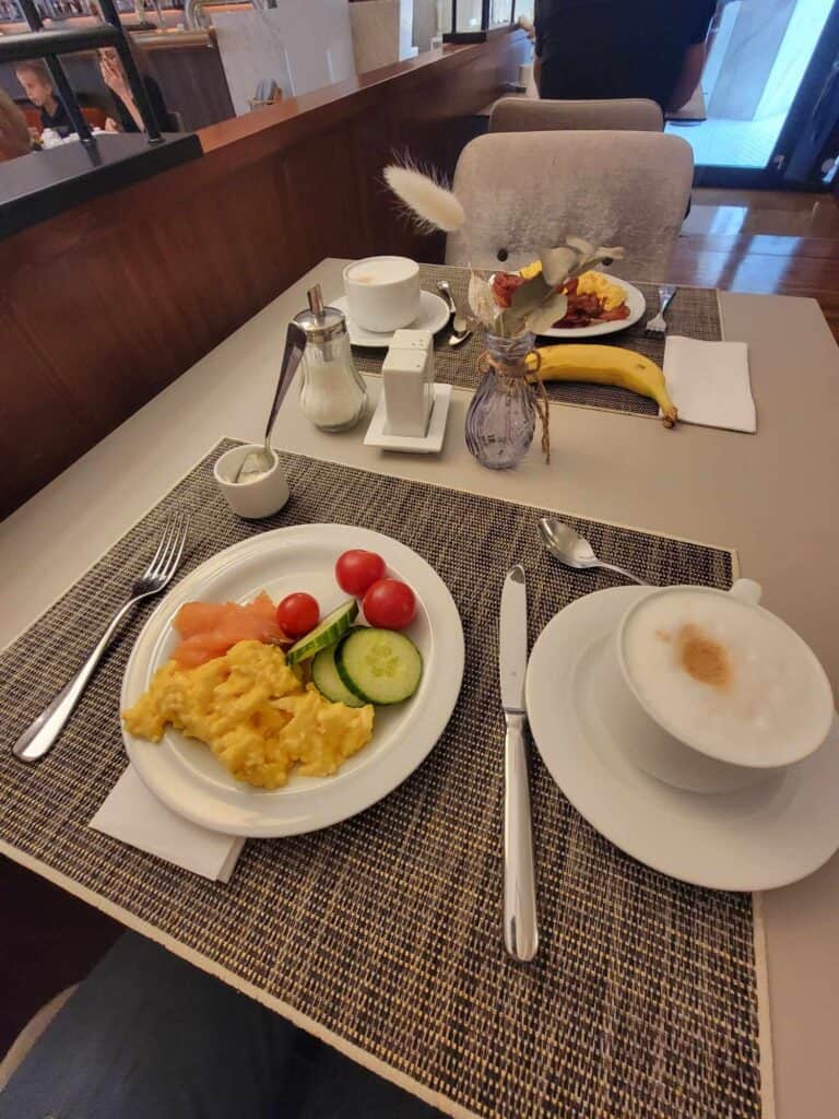 Two plates of breakfast, one with scrambled egss, lox, cucumber and tomato with a cappuccino 