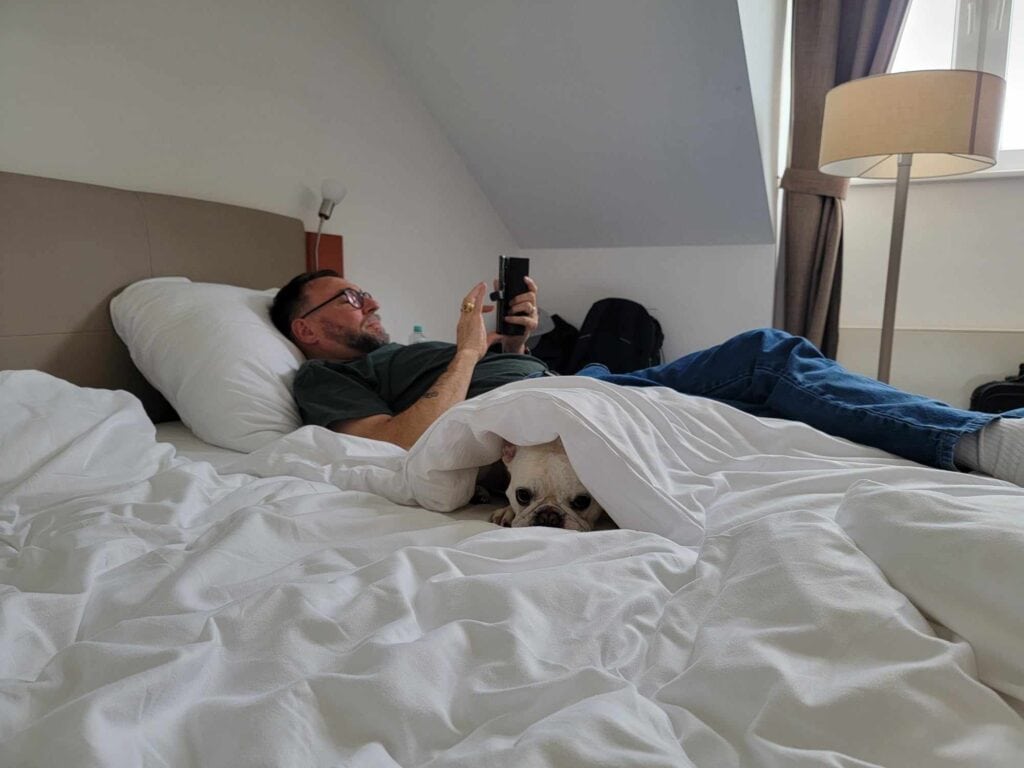 A man lies on a bed with white linens, and a french bulldog peeking out from under the covers