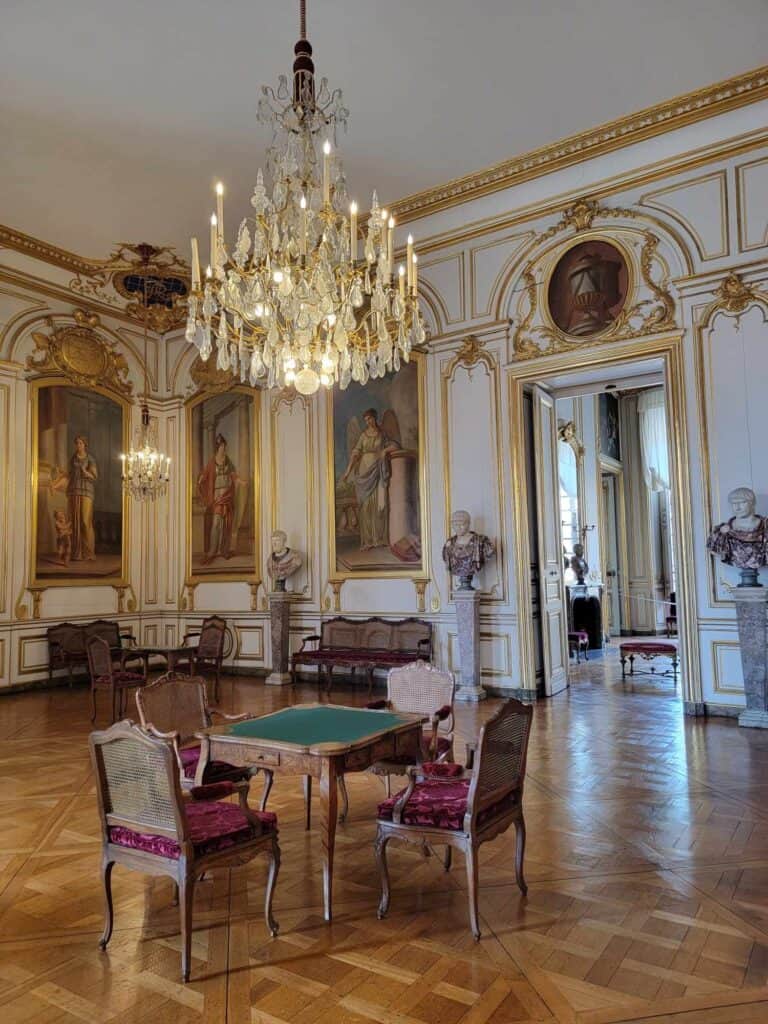 A room in Palace Rohan with a crystal chandelier, a card table and chairs, parquet floors, large paintings, and gold detailing everywhere in the room