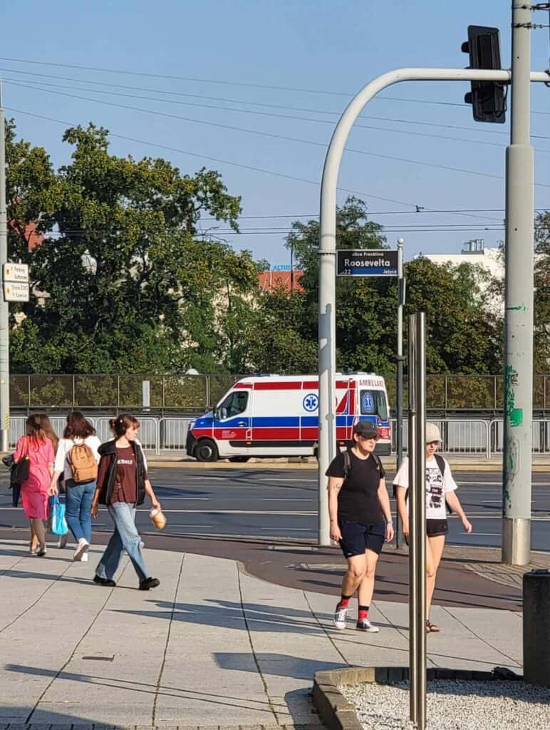 People walking on the sidewalk and a white ambulance with red and blue stripes drives behind on the street in Poznan